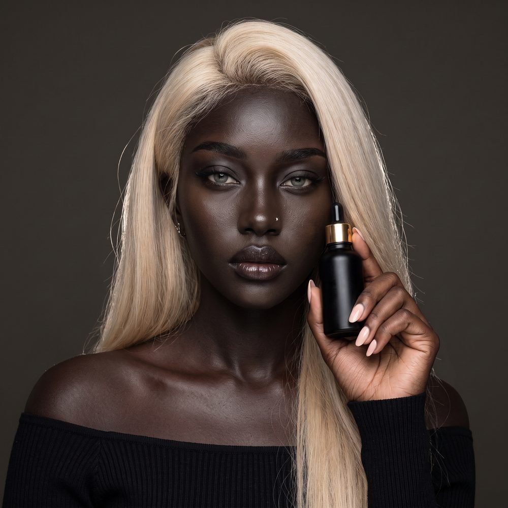 Aïra tenant un flacon compte-gouttes noir au bouchon doré, visuel de type campagne beauté